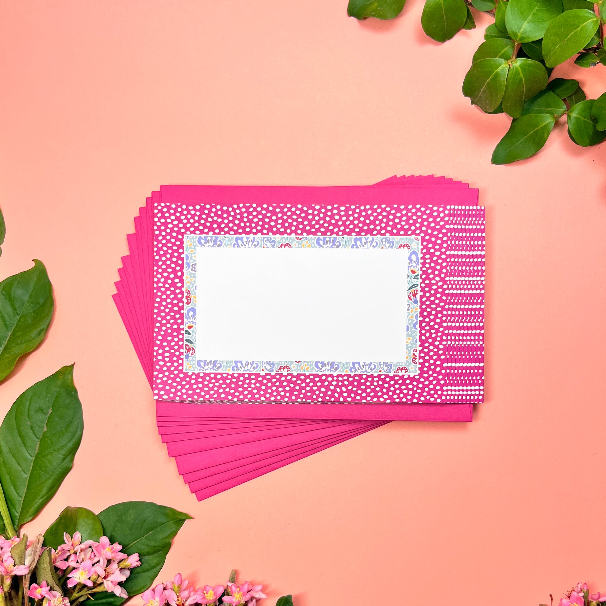 Stack of pink greeting cards with a decorative card on top, surrounded by green leaves and flowers on a pink background